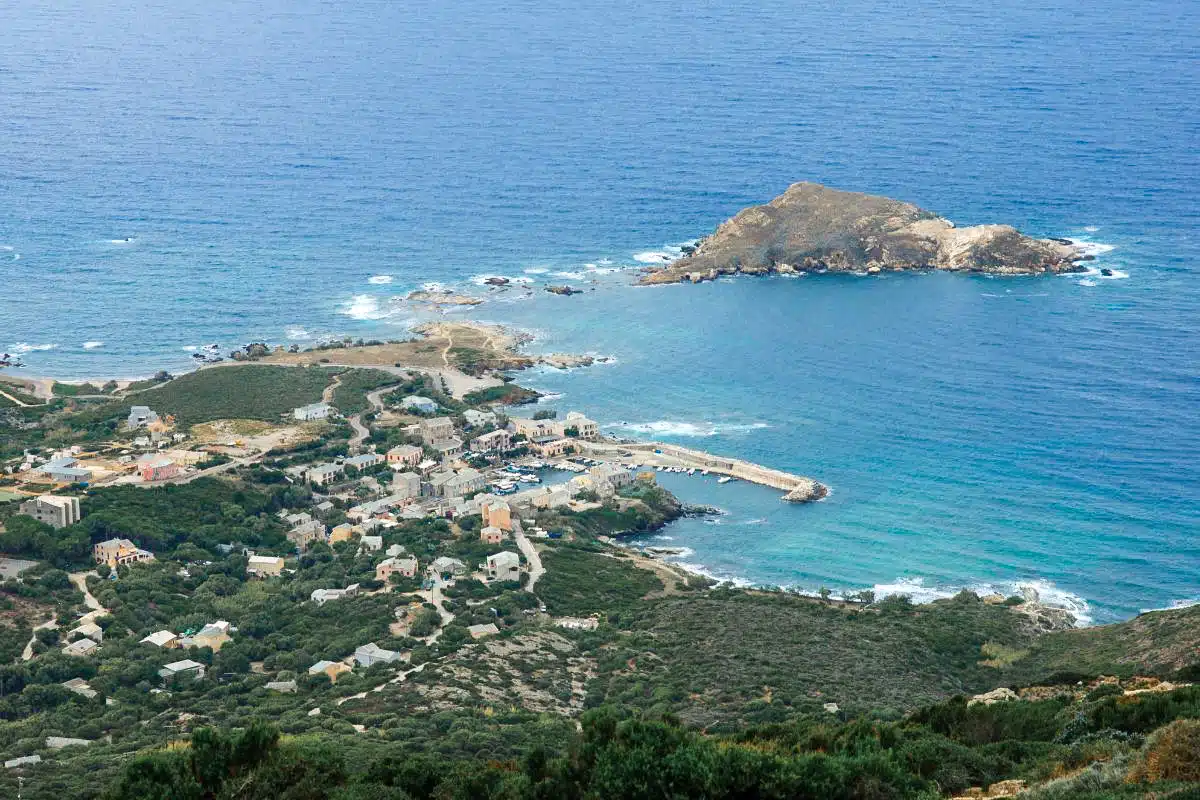 An aerial view of a coastal village with scattered buildings, green vegetation, and a blue sea, with a rocky islet close to the shore.