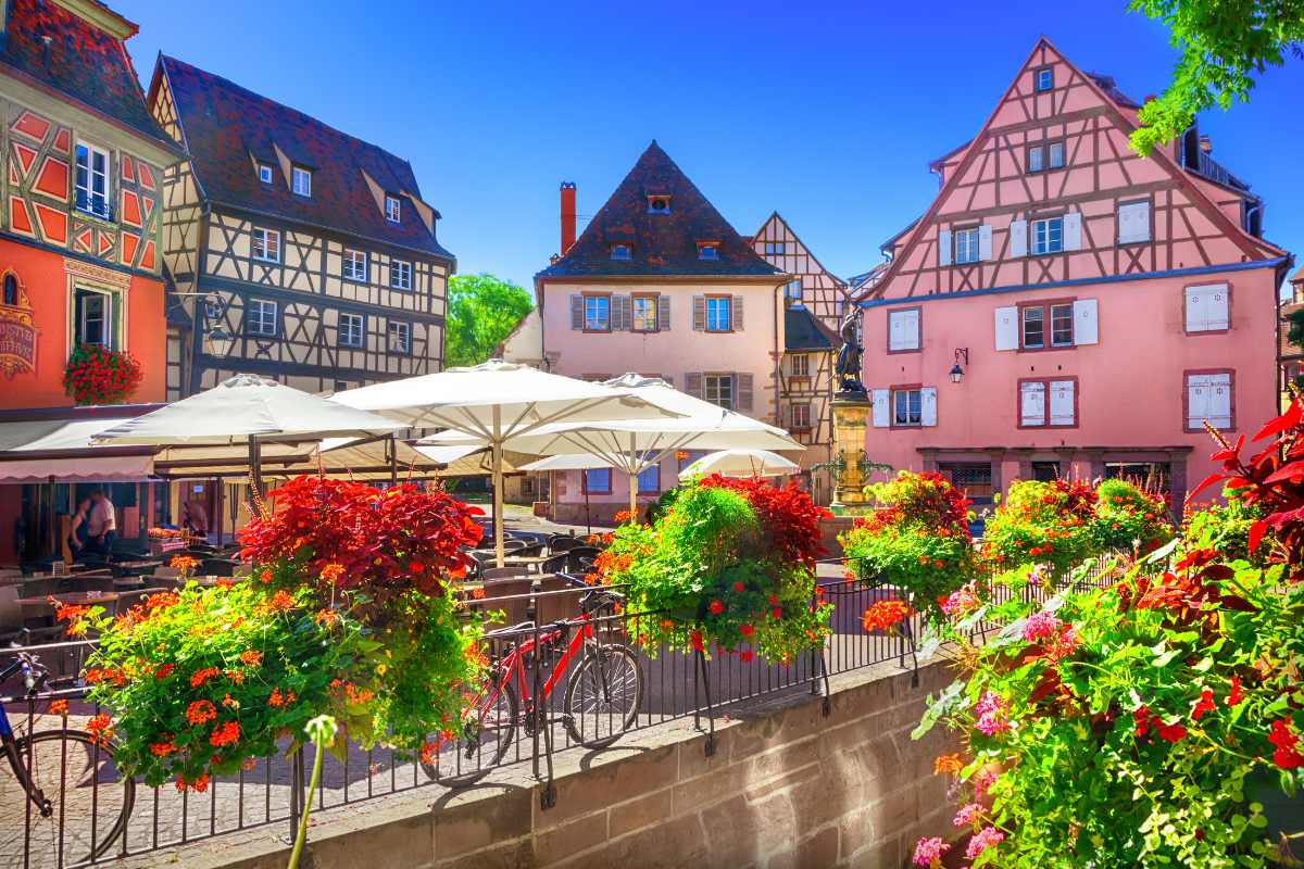 A bright, sunny day in an Alsace village, where colorful half-timbered buildings frame a lively square with outdoor café umbrellas, vibrant flowers, and a bicycle leaning against a railing, inviting a leisurely day out.