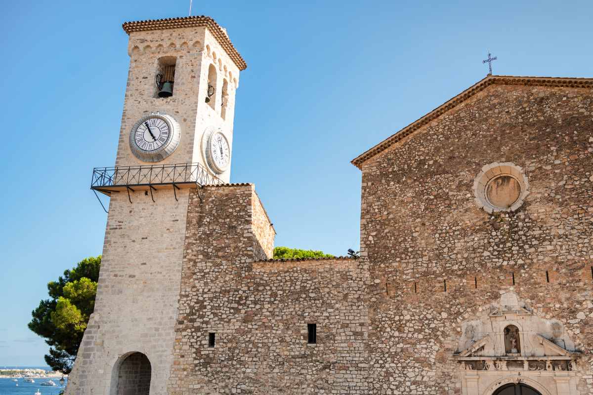 The historic clock tower of the Notre-Dame d'Espérance church standing tall against a clear blue sky in the old quarter of Cannes, with the fortified walls of the medieval building visible.