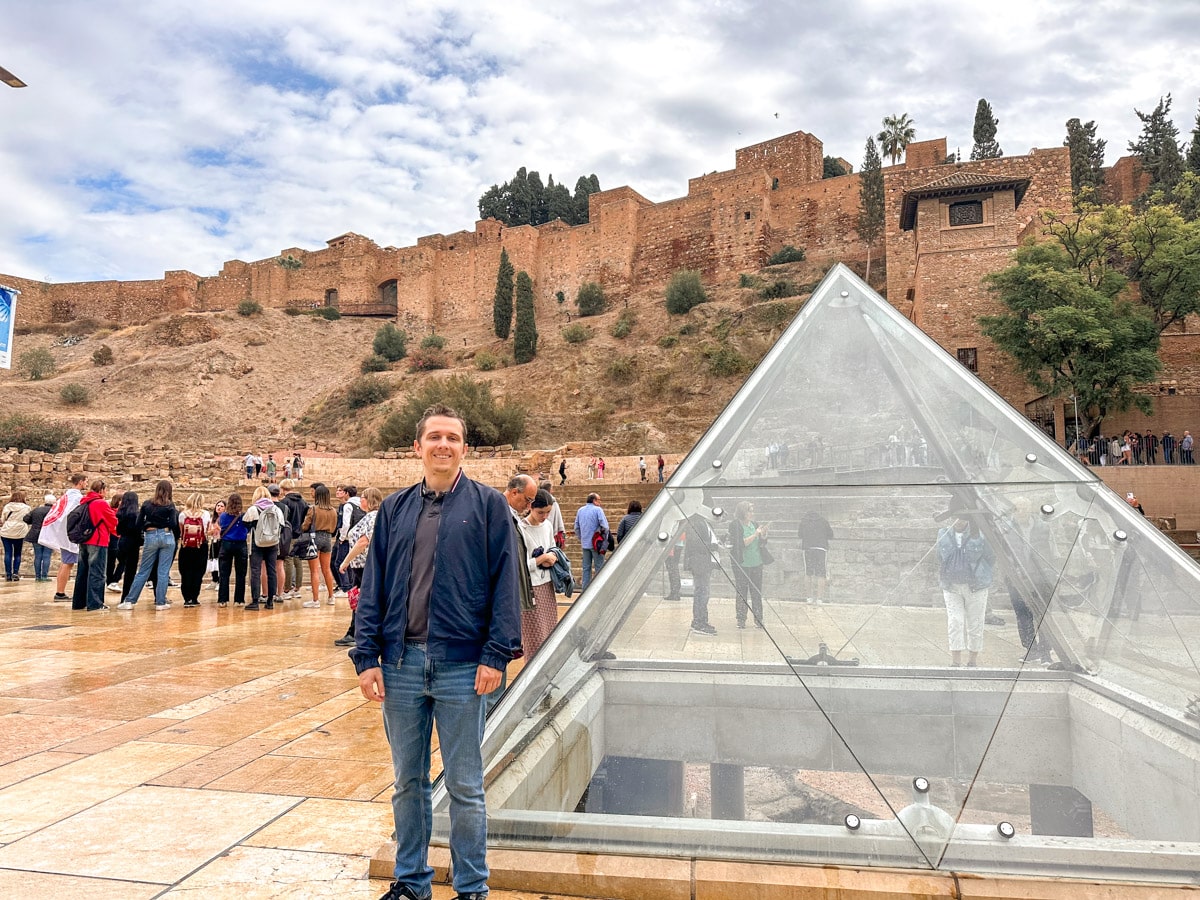 A man smiling in front of the reflective glass pyramid at the Alcazaba entrance, with the fortress walls and visitors in the background. It is the husband of the author. 