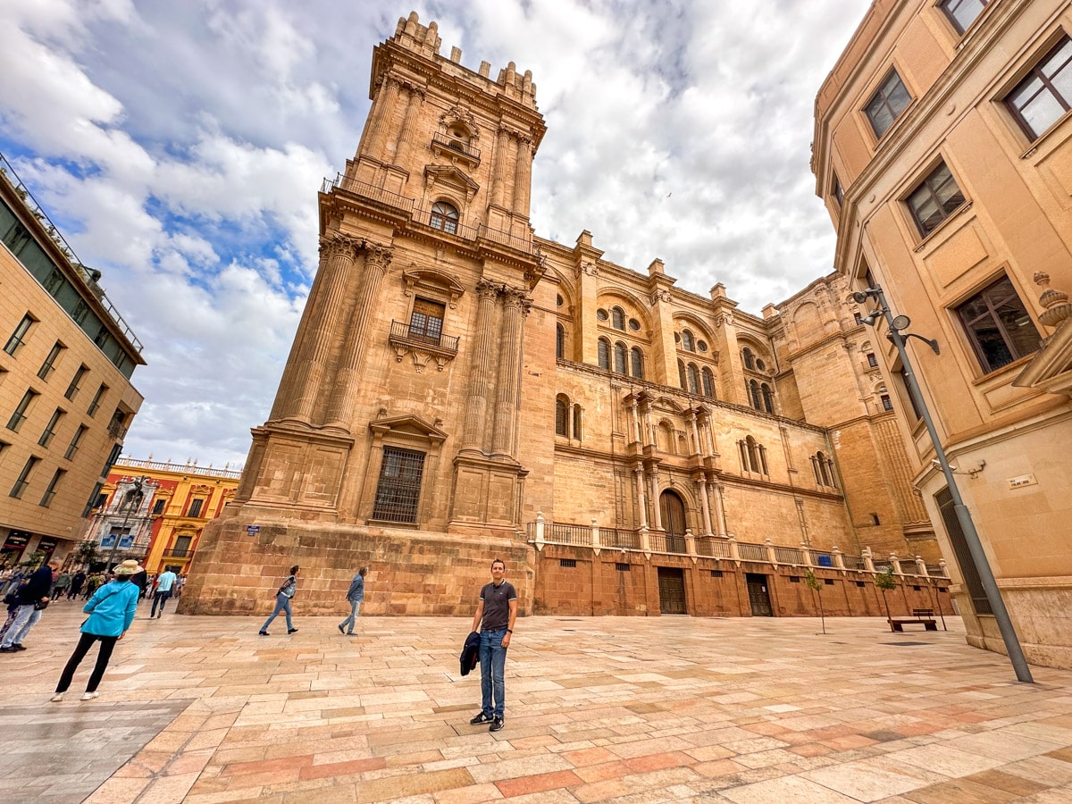 The towering presence of Málaga Cathedral's side façade, seen from a square where people are walking and relaxing. you can see the author's husband in the picture as well enjoyting his 1 day in malaga. 