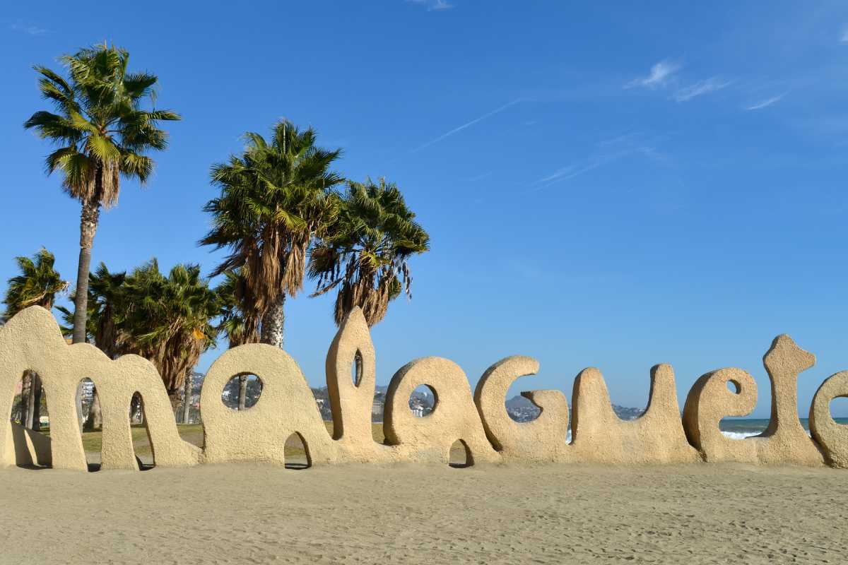 Playa la Malagueta beach in Malaga with some palm trees and the ocean behind the big signs saying malagueta in sand. 