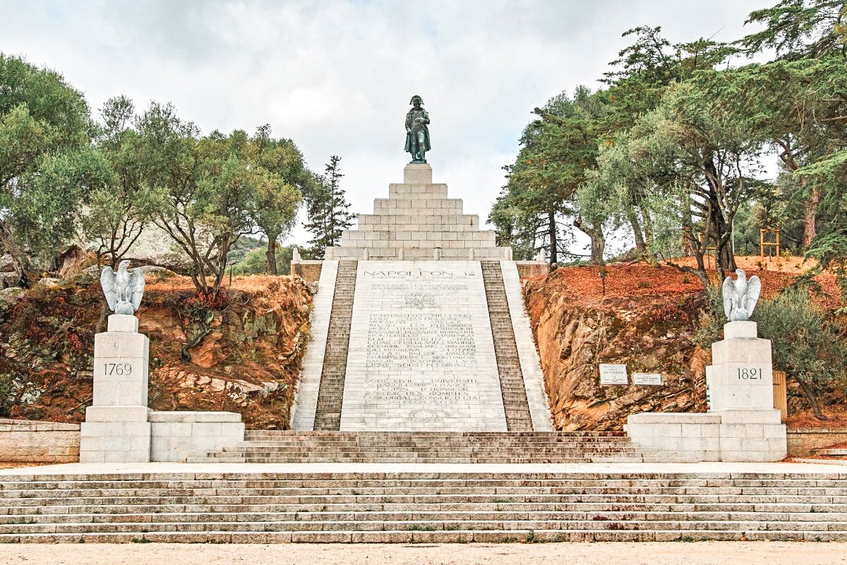 A stone monument to Napoleon Bonaparte, with a statue standing atop a series of steps, flanked by sculptures of eagles and inscriptions, with trees and a cloudy sky in the background. Totally worth visiting in one day in Corsica.