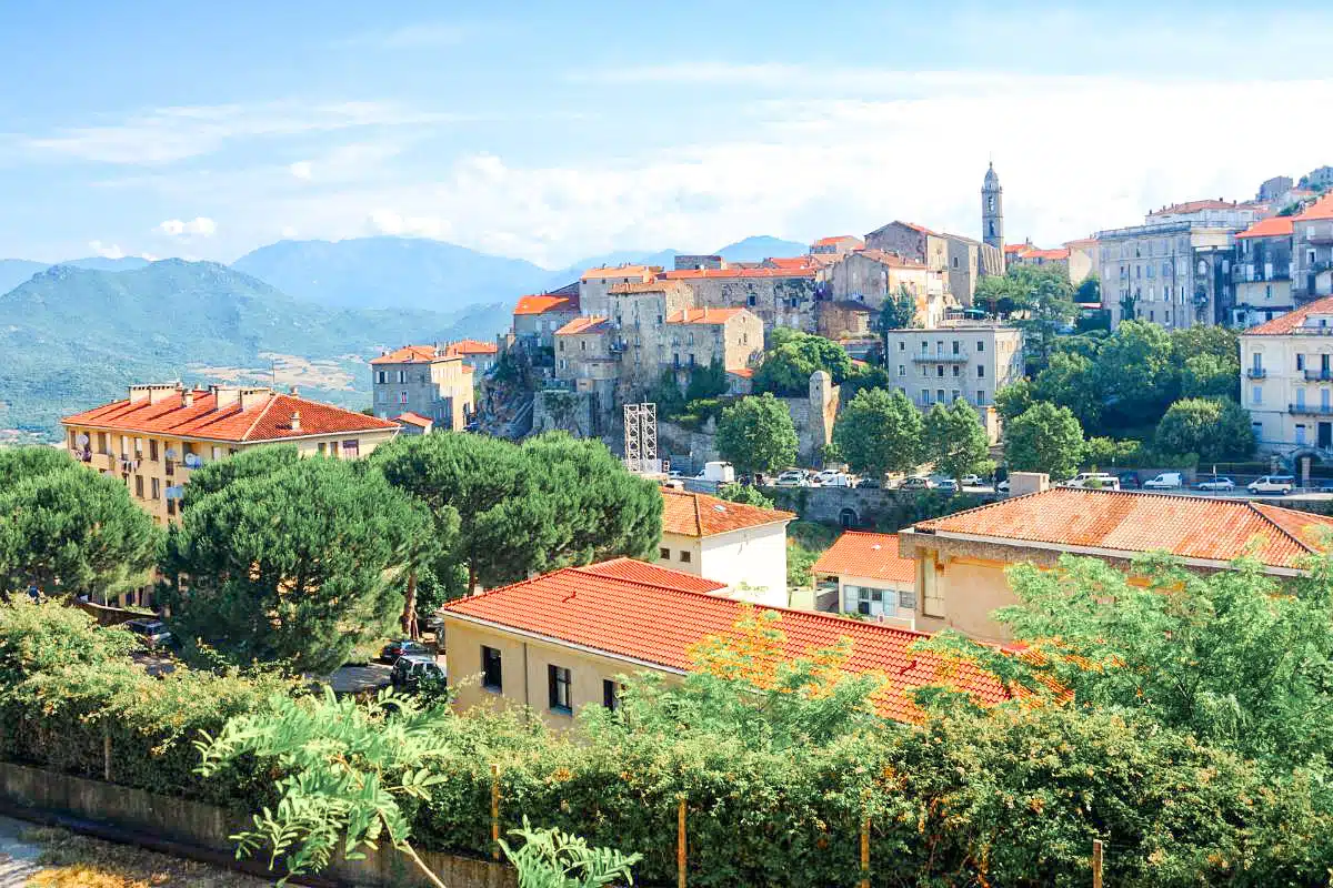 An old hilltop town with stone buildings and a church, surrounded by lush greenery and mountains in the backdrop, under a partly cloudy sky.