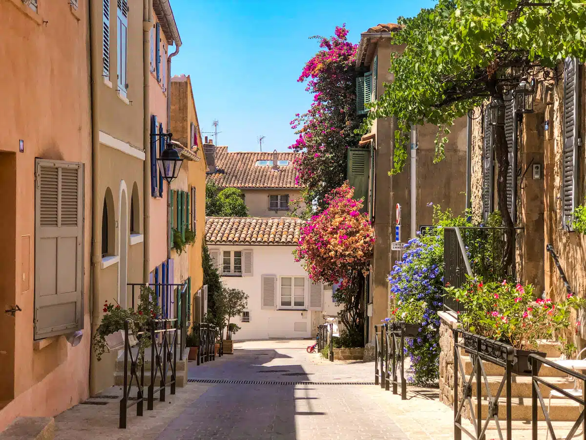 A charming, narrow street in the old town of Saint Tropez, lined with traditional pastel-colored houses adorned with flowering plants and shutters, evoking a peaceful, picturesque setting.