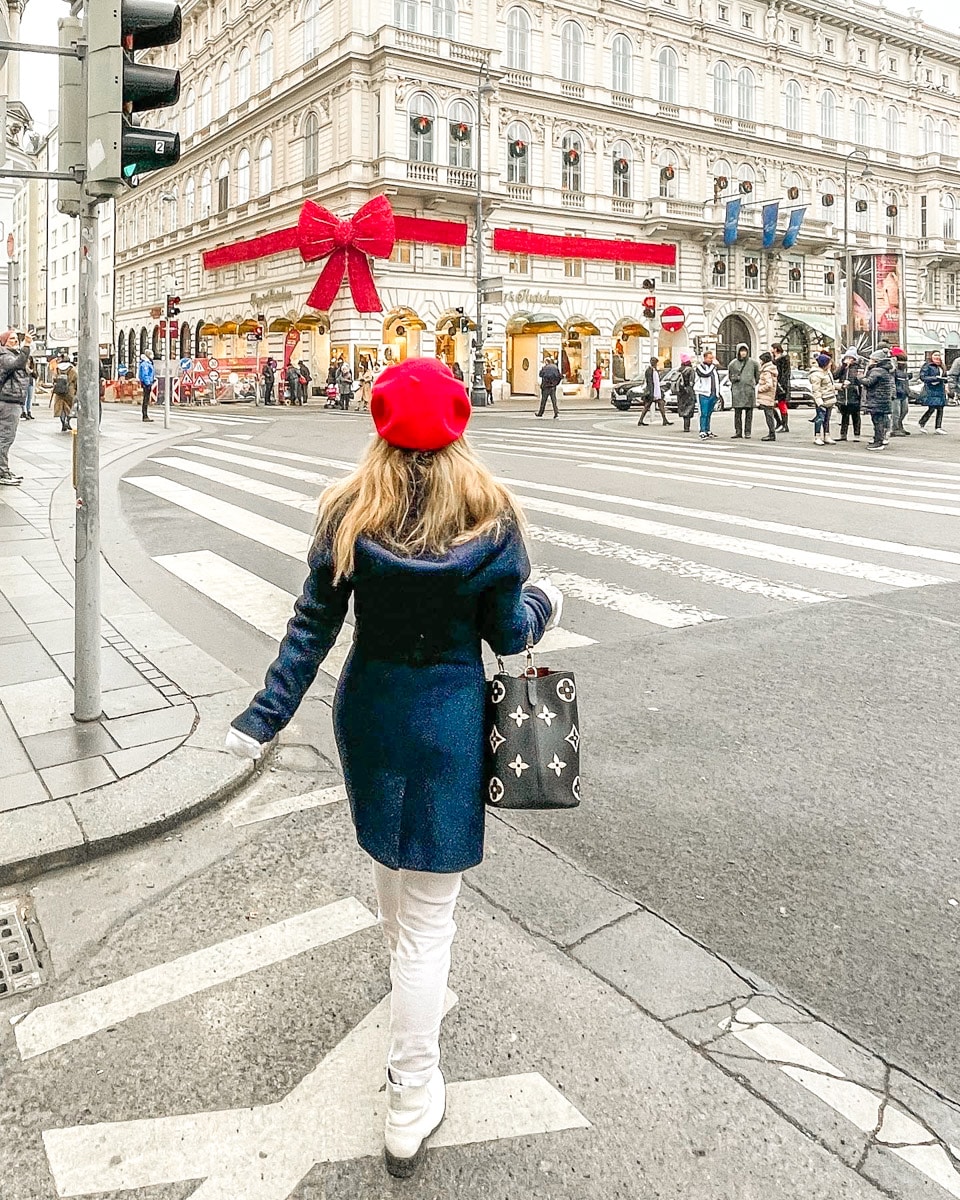 the author in a red hat and blue coat, seen from behind, walking across the street towards a festively decorated building with a big red bow.