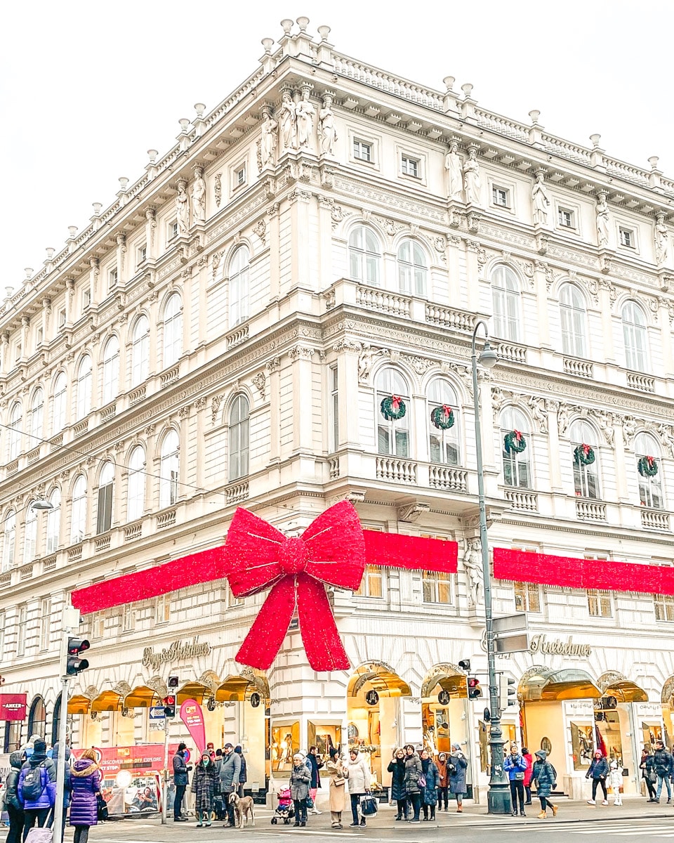 "A grandiose building with intricate architectural details and statues, adorned with a huge red bow, signaling festive times, as people wander around on a busy street. This picture shows perfectly why winter is the best time to visit vienna.