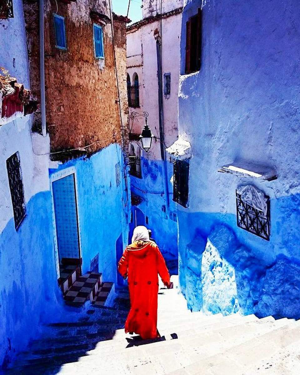 A lone figure in a vibrant orange garment walking down a narrow blue alleyway in Chefchaouen, with sunlight casting deep shadows, creating a striking contrast of colors