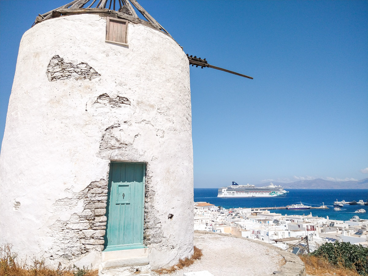 A traditional white windmill standing solitary atop a hill in Mykonos, overlooking a panoramic view of the town and harbor with cruise ships in the distance, capturing the blend of old and new