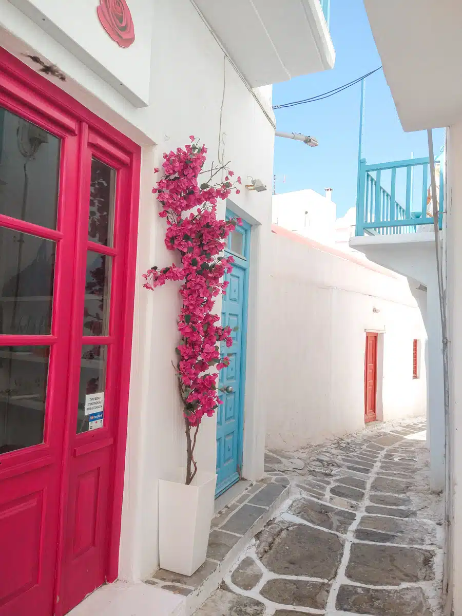 A charming street in Mykonos, adorned with vibrant pink bougainvillea, contrasting against white-washed walls and colorful doors, embodying the island's picturesque aesthetic.