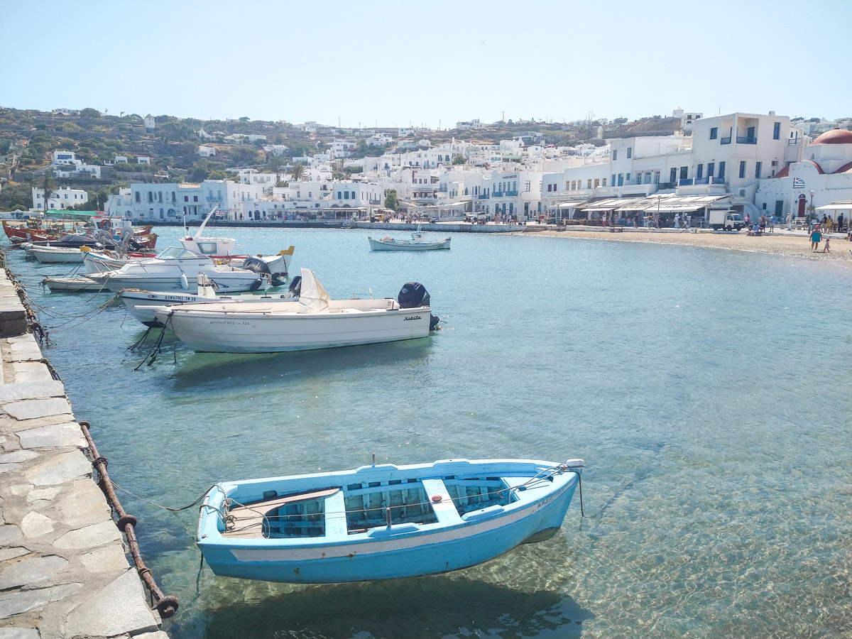 A peaceful harbor scene in Mykonos, with small boats floating on the clear water and the town's buildings in the distance, portraying the island's maritime culture. This spot in Mykonos is definitely worth visiting