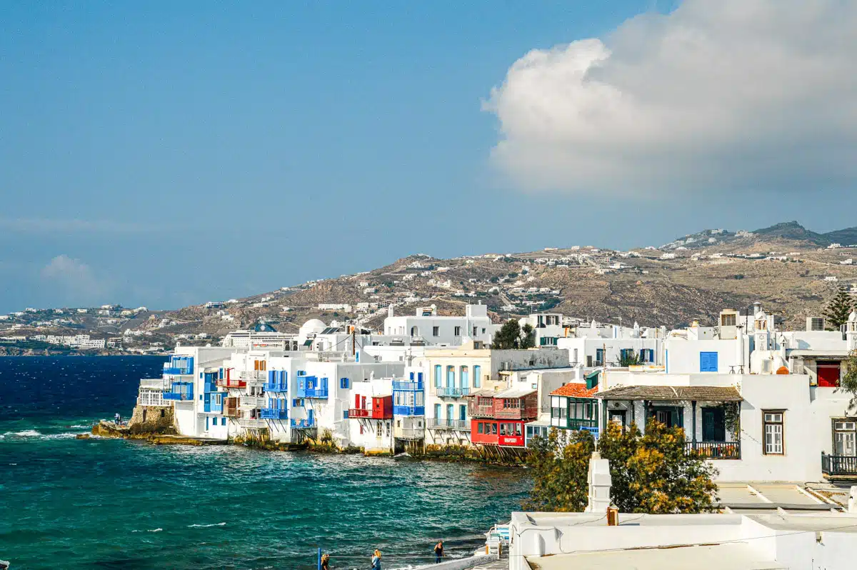 A coastal view of Mykonos with the sea in the foreground, showing a mix of traditional and modern architecture along the shore, with a cruise ship anchored in the background, representing the island's appeal as a top tourist destination.