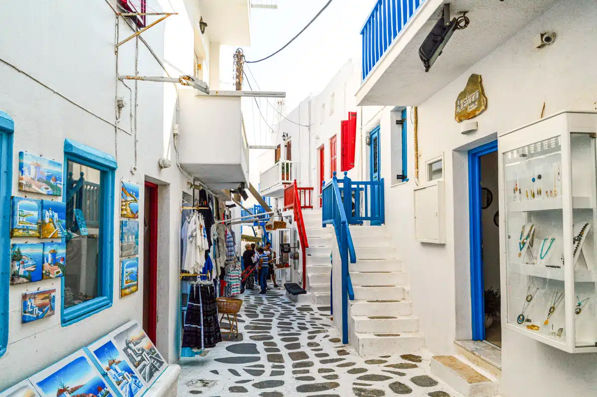 A narrow alley in Mykonos town, flanked by white buildings with colorful doors and balconies, with shops displaying art and souvenirs, capturing the bustling atmosphere of a Greek island street.