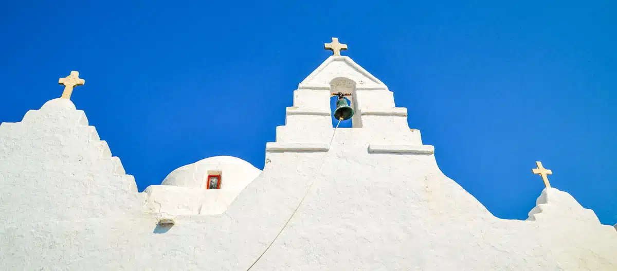 The iconic façade of a white Mykonian church with three bells, set against a clear blue sky, symbolizing the religious heritage and architectural simplicity of the Cycladic island
