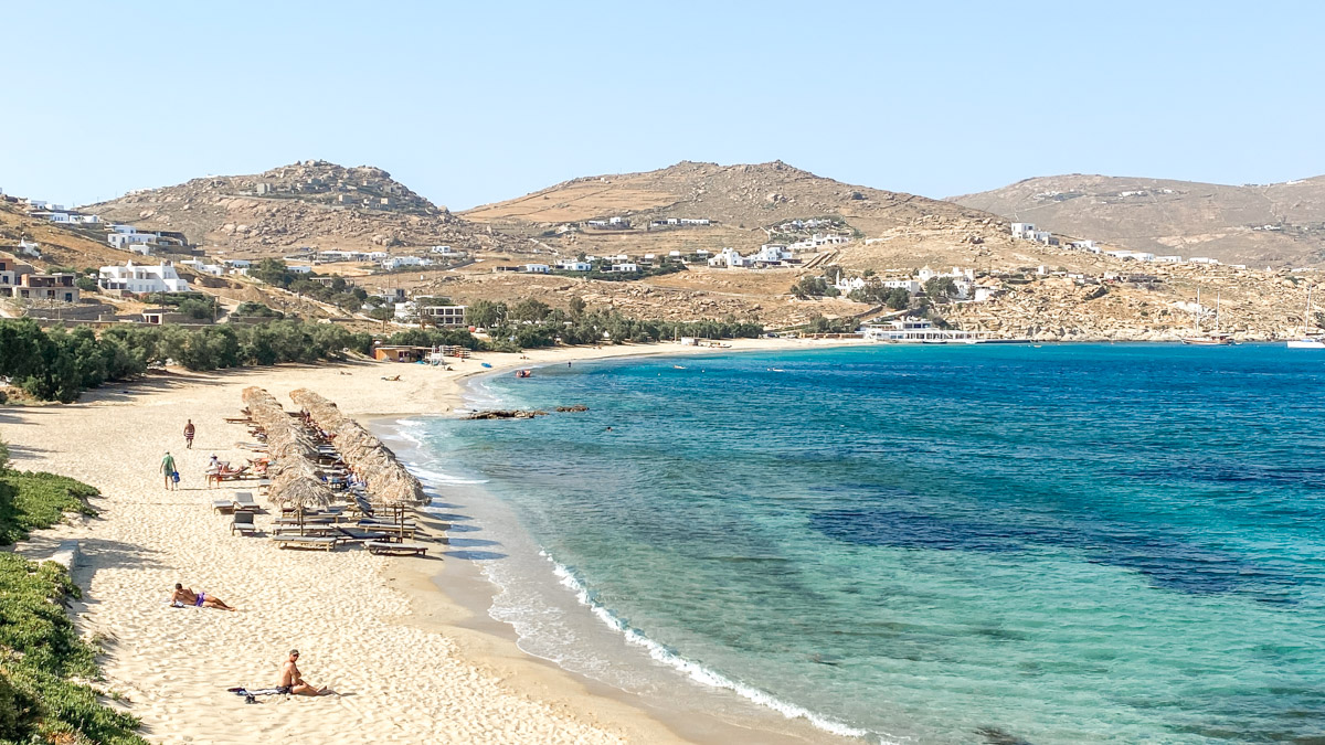 A sun-drenched beach on the coast of Mykonos, featuring golden sands, beach loungers under thatched umbrellas, and the azure Aegean Sea, with the island's rugged hills and whitewashed buildings in the background.