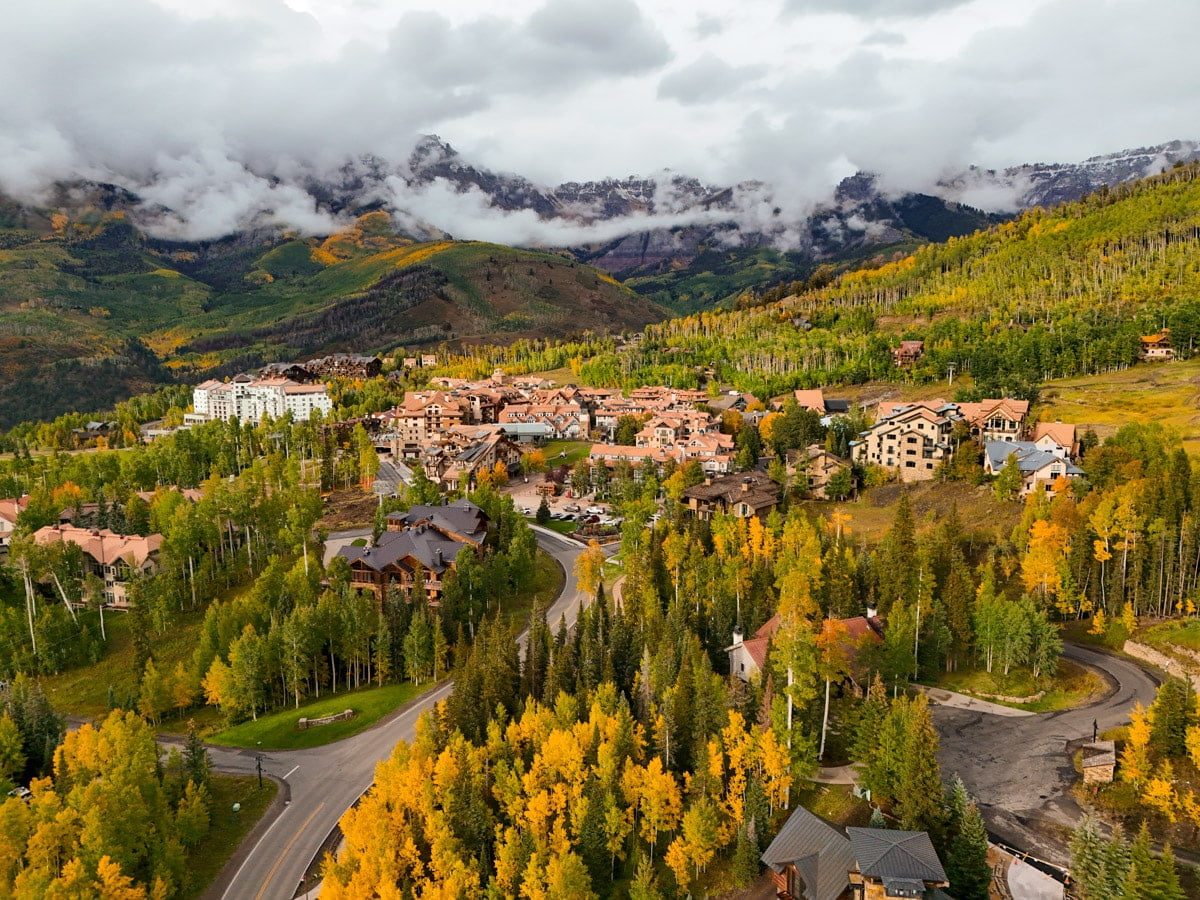 High-angle view of Telluride with residential buildings amidst vibrant yellow and green aspen forests, with mountains partially shrouded in clouds in the background.