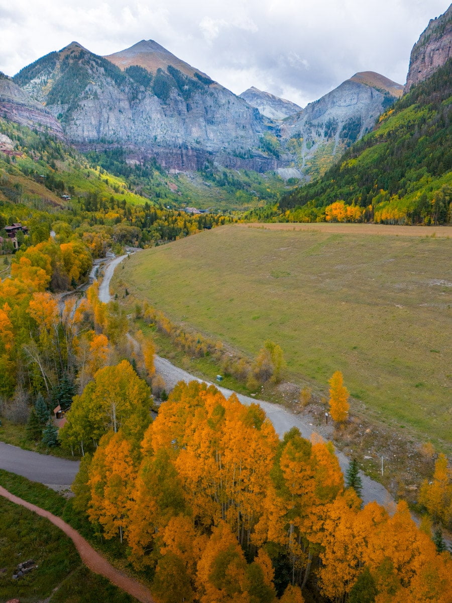 Aerial shot of a valley in Telluride, featuring a river, a meadow, and colorful aspen trees with rugged mountains in the distance.