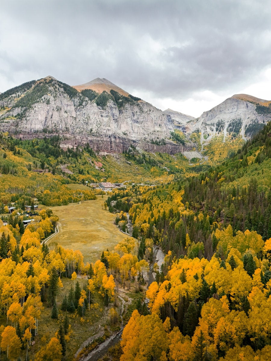 Aerial shot of a valley in Telluride, featuring a river, a meadow, and colorful aspen trees with rugged mountains in the distance.
