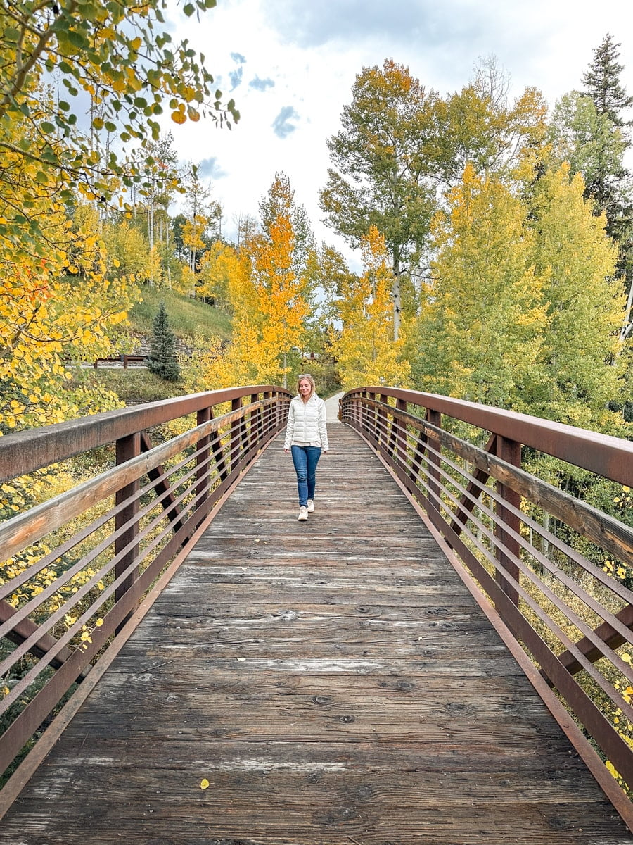 Photo of a woman walking on a wooden bridge surrounded by vibrant yellow aspen trees in fall. It is the authors wife in typical fall fashion.