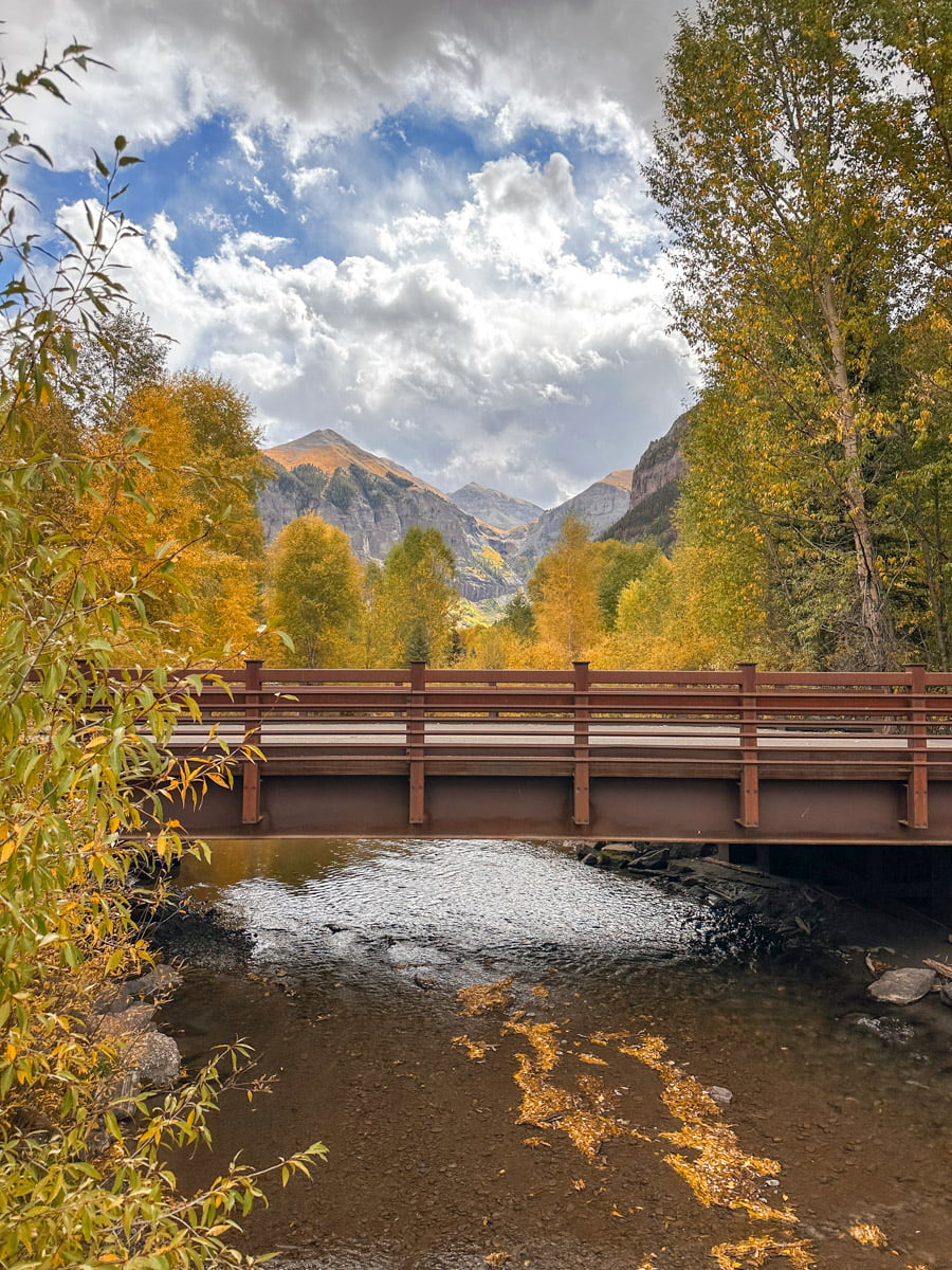 A wooden bridge over a creek surrounded by golden aspen trees and a mountain backdrop. You can see this beautiful view in Telluride in the fall.