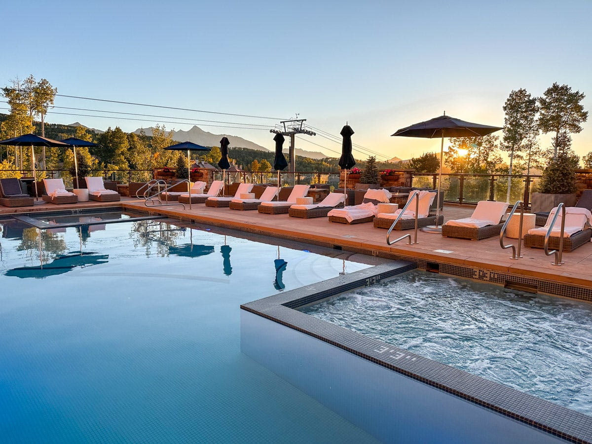 Poolside view at sunset with lounge chairs and a reflective blue pool, surrounded by autumn-colored trees and mountains.