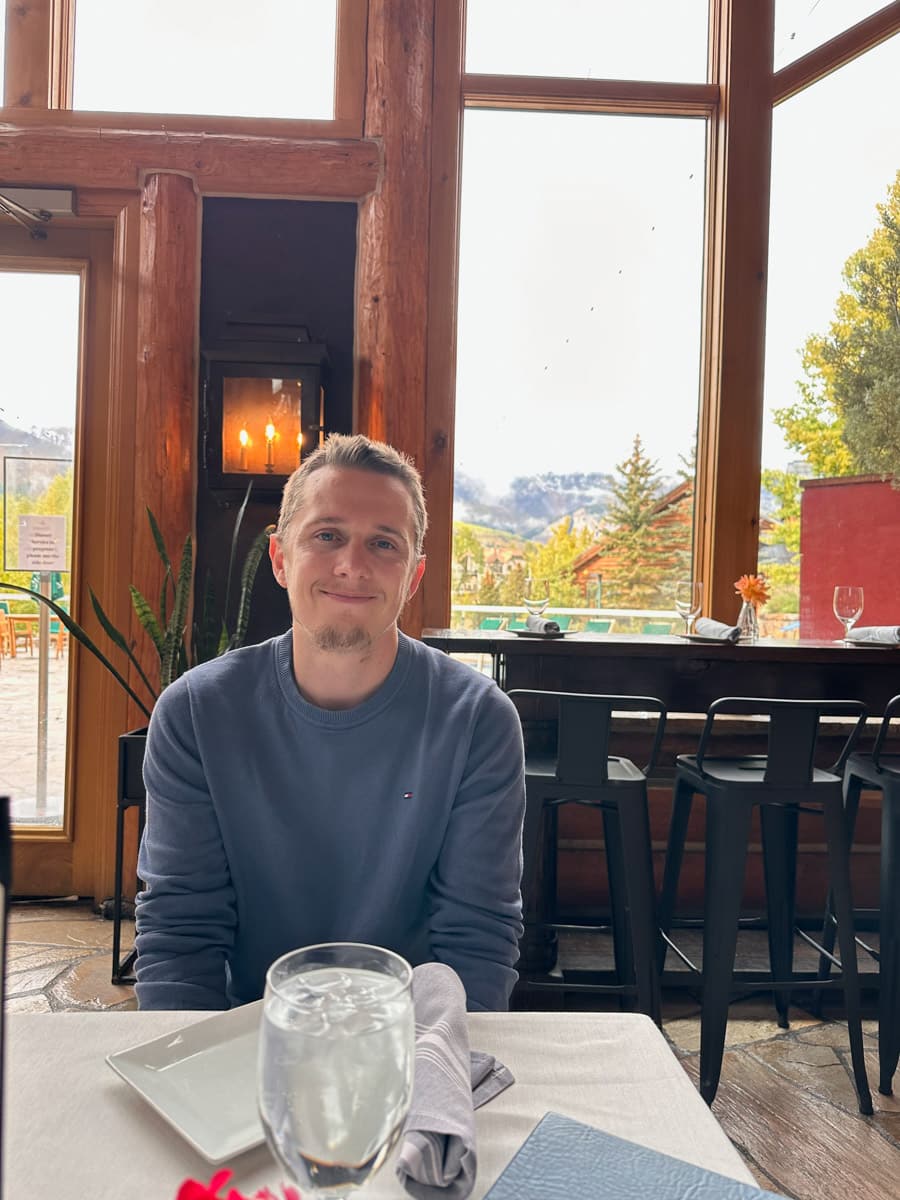 The author sitting at a restaurant table, smiling, with a scenic view through the window.