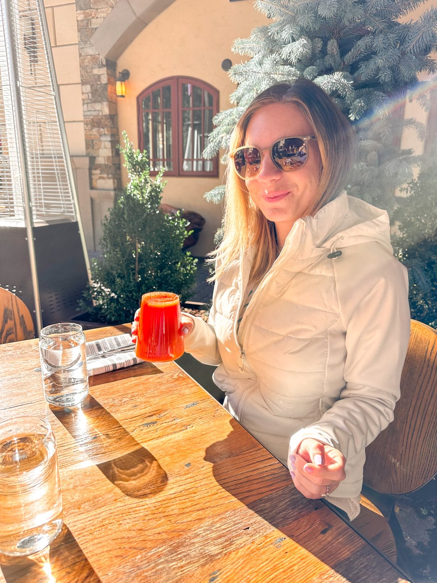 A woman sitting at an outdoor restaurant table, holding a red drink, with autumn scenery in the background.