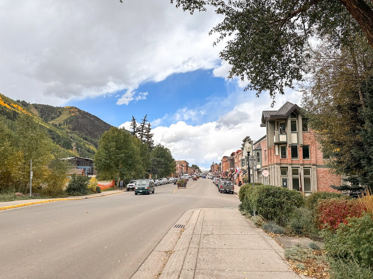 A wide street view in Telluride, lined with historic buildings and autumn-colored trees, with mountains in the distance.