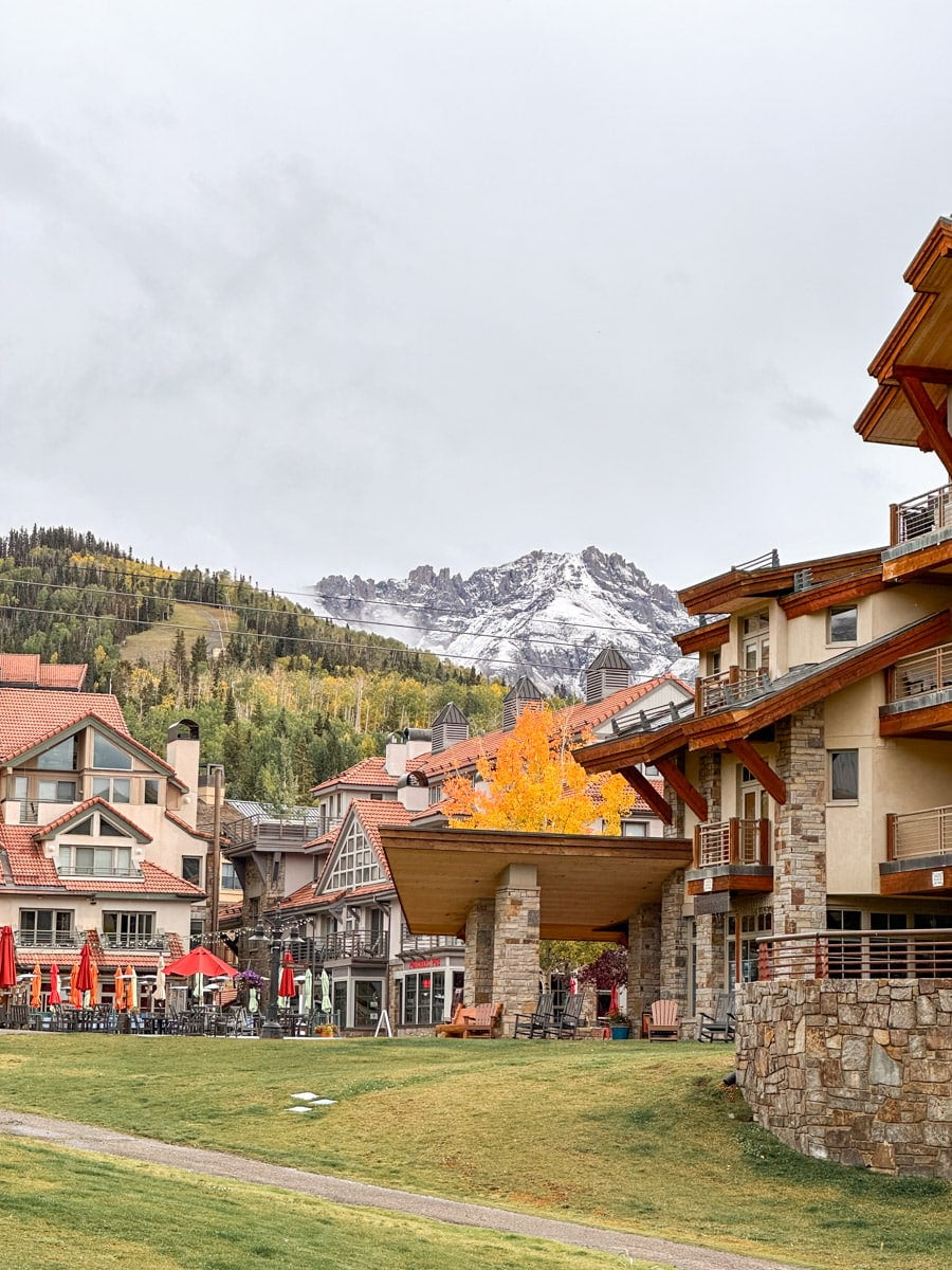 A view of a resort-style outdoor area with buildings and autumn trees against a backdrop of snow-capped mountains.