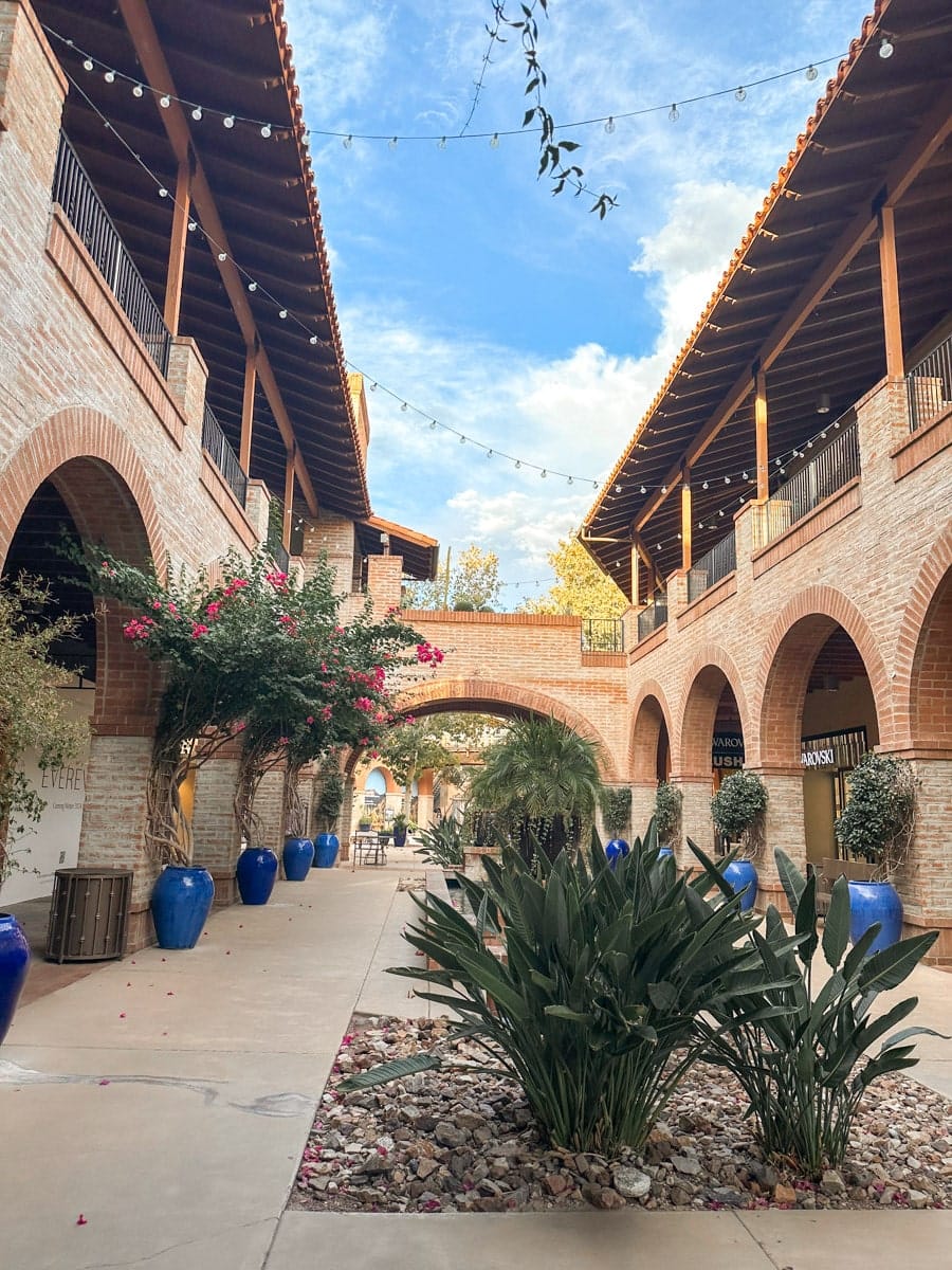 A charming courtyard with Southwestern architecture featuring terracotta tiles, arched walkways, and vibrant blue planters under strings of lights. La Encantada Shopping Center in tucson