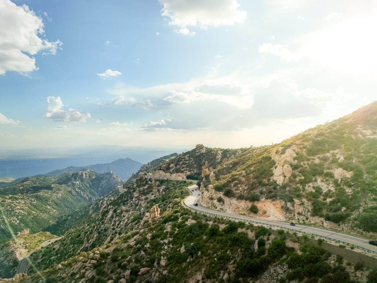 Aerial view of a winding mountain road at Mount Lemmon, showcasing lush greenery and distant mountains under a partly cloudy sky.