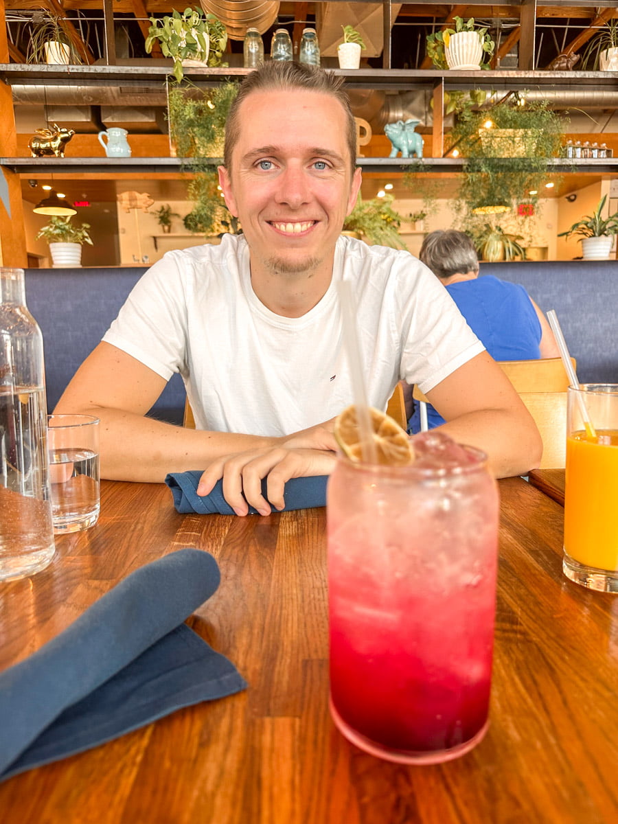 A young man smiling at the camera while sitting at a restaurant table, with a vibrant pink drink in front of him and a modern decor background