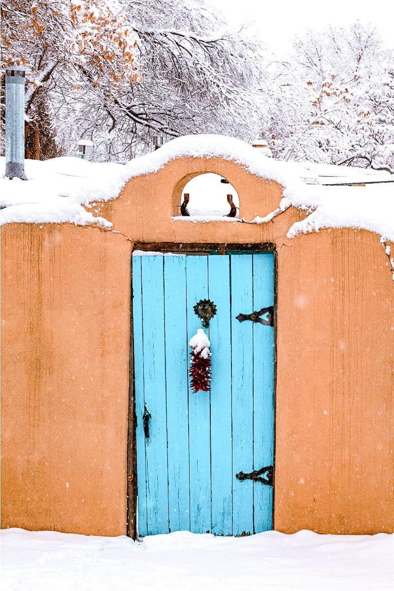 stunning red adobe house with blue door covered in snow in santa fe