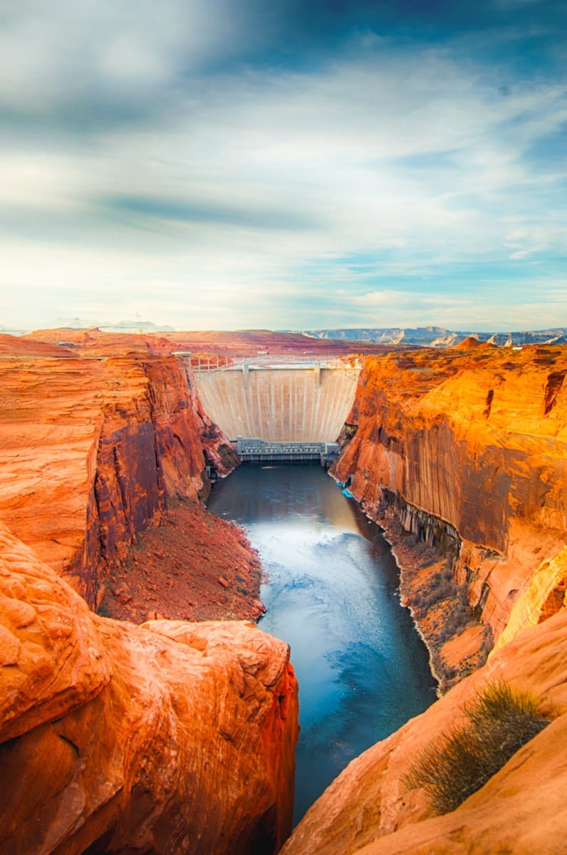 The towering Glen Canyon Dam holding back the Colorado River, framed by the red rock cliffs of the canyon, under a partly cloudy sky