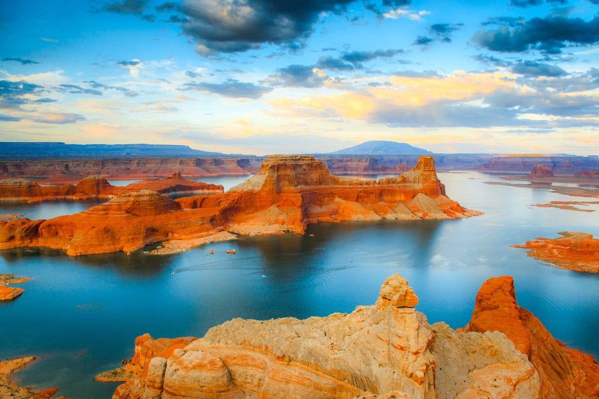 A stunning view of Lake Powell, surrounded by red rock formations and mesas, with the blue water reflecting the golden light of sunset.