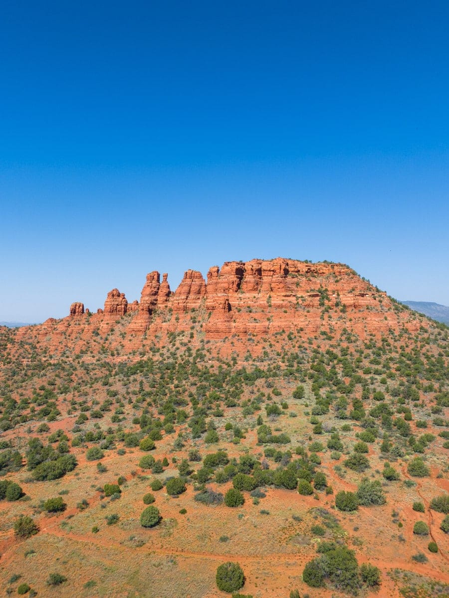 A distant view of red rock formations in Sedona under a clear blue sky with green desert shrubs dotting the landscape.