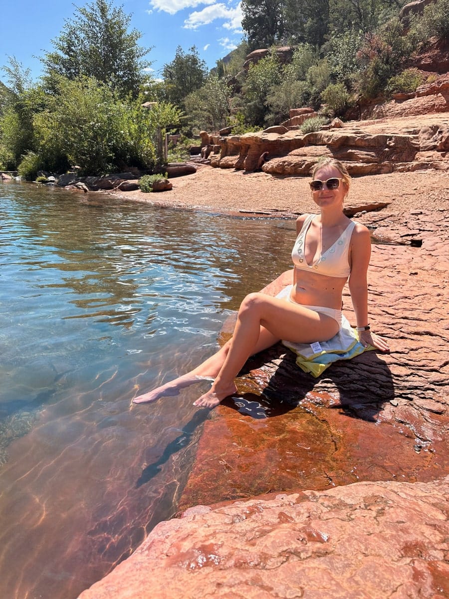 the author's wife relaxing by the water at Oak Creek in Sedona, Arizona, sitting on the smooth red rocks under a sunny sky.