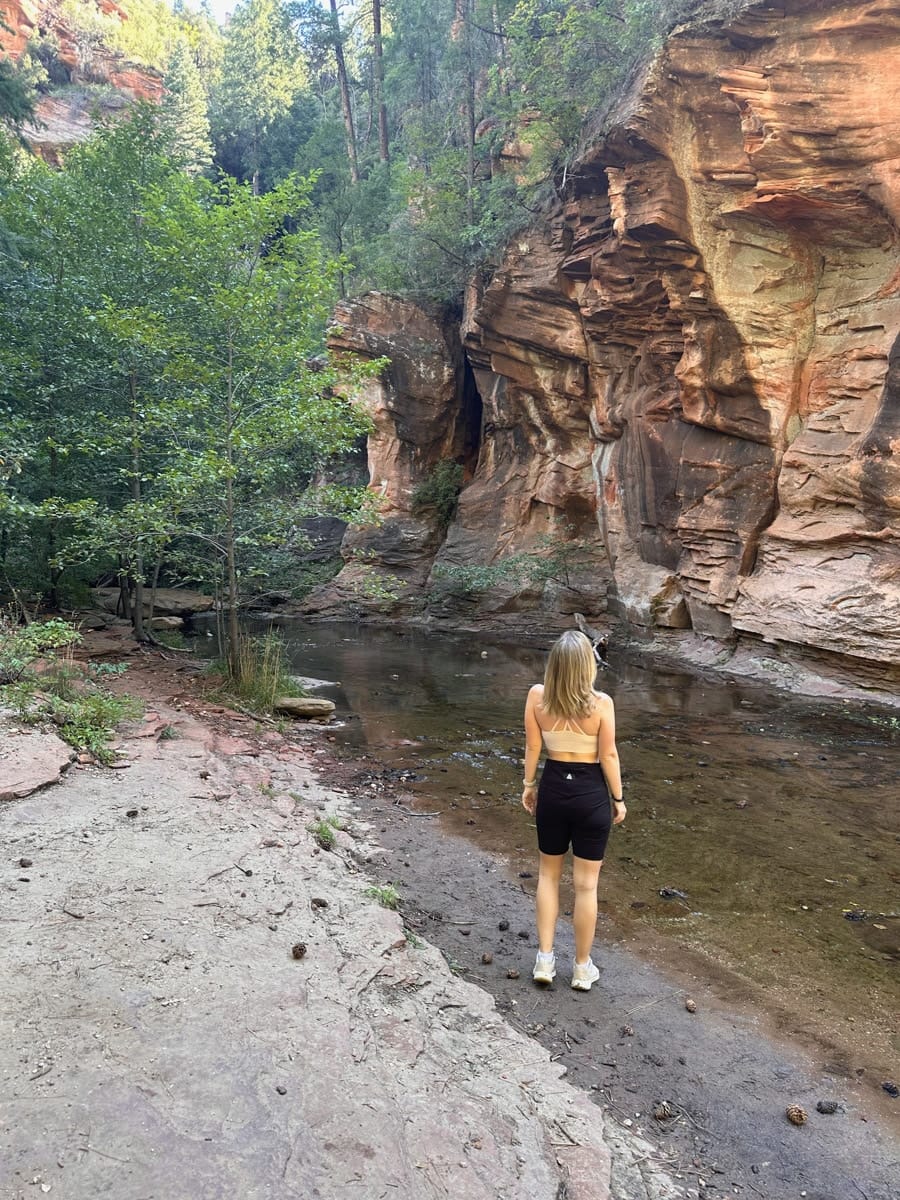 the author's wife standing beside a calm stream in a peaceful canyon with red rock walls and lush greenery in Sedona, Arizona.