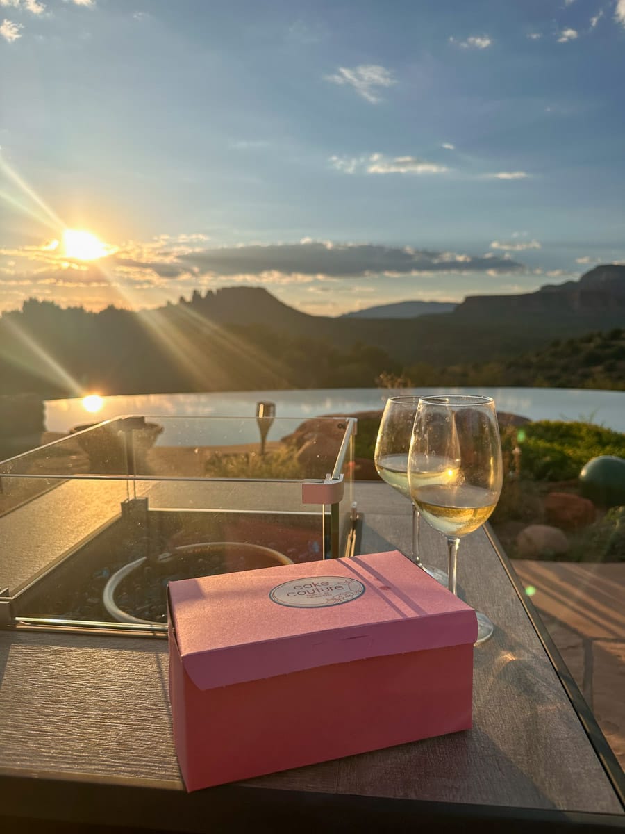Two wine glasses on a patio table with a pink dessert box, overlooking a stunning sunset view of Sedona's red rocks.