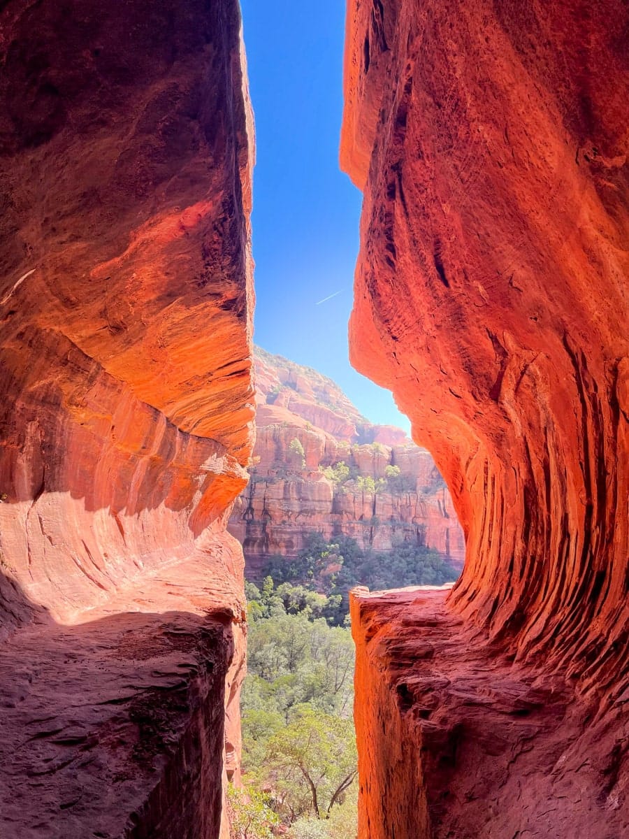 View from within a narrow red rock cave in Sedona, Arizona, with a bright blue sky visible between the canyon walls.