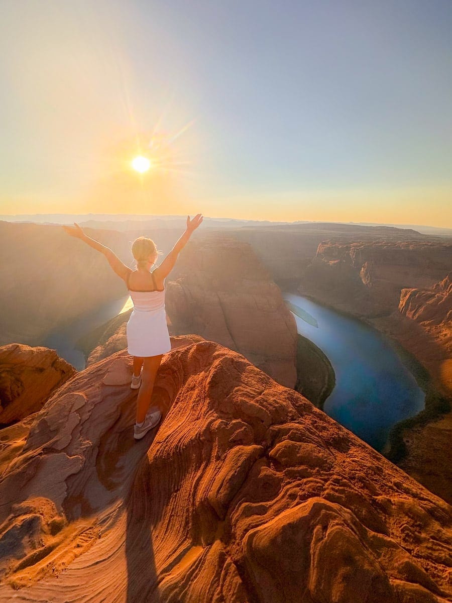 the authorin a white dress standing with arms raised, enjoying the stunning sunset view at Horseshoe Bend in Page, Arizona, with the Colorado River and red rock formations in the background.