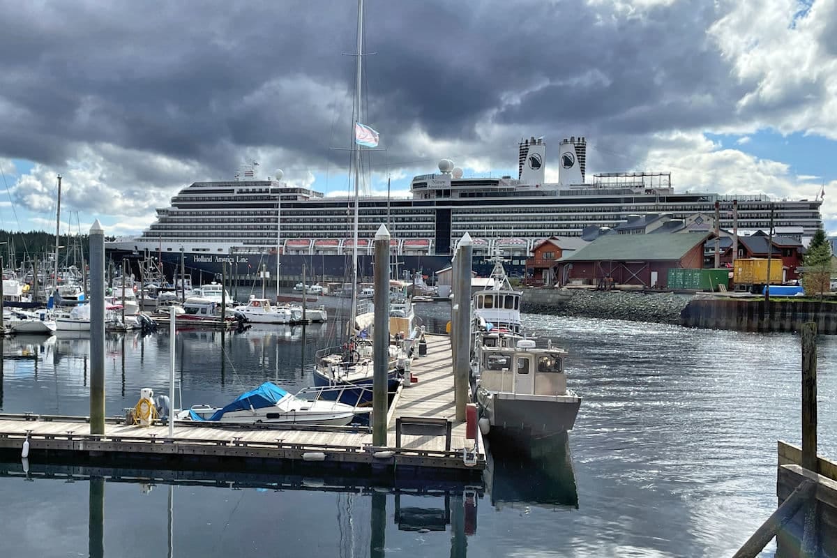 A Holland America cruise ship docked at a marina in Sitka, Alaska, surrounded by smaller boats and a cloudy sky.