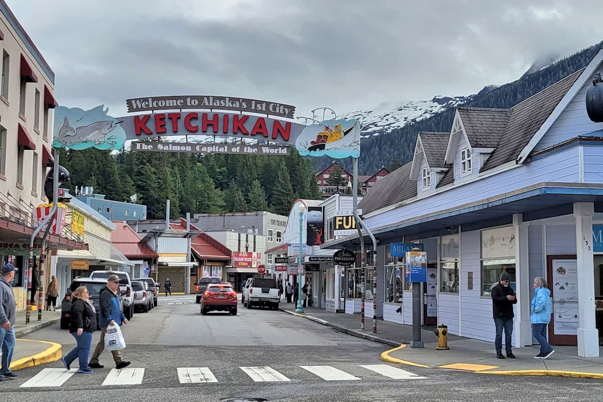 The famous 'Welcome to Ketchikan, Alaska's 1st City' sign arching over a downtown street, surrounded by shops, cars, and pedestrians in the Salmon Capital of the World.