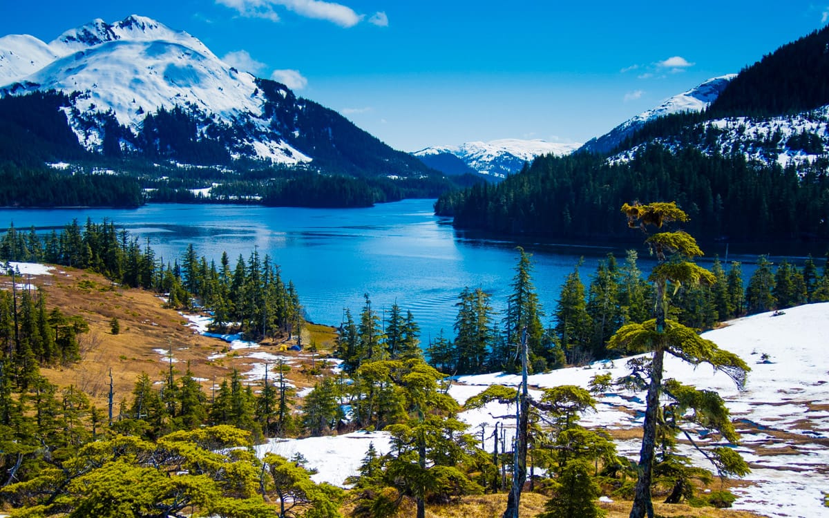 Snow melts slowly from the mountains as spring approaches at the end of May in south-central Alaska. An Alaskan wilderness scene featuring a serene blue lake surrounded by snowy mountains and dense evergreen trees under a bright blue sky.