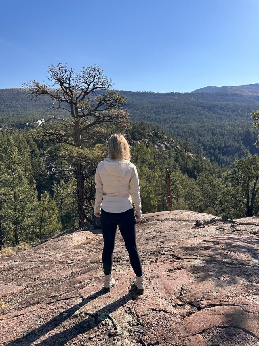 The author's wife standing on a rocky overlook surrounded by a forest of green pines, gazing out at a peaceful and vast wilderness under a clear, sunny sky.