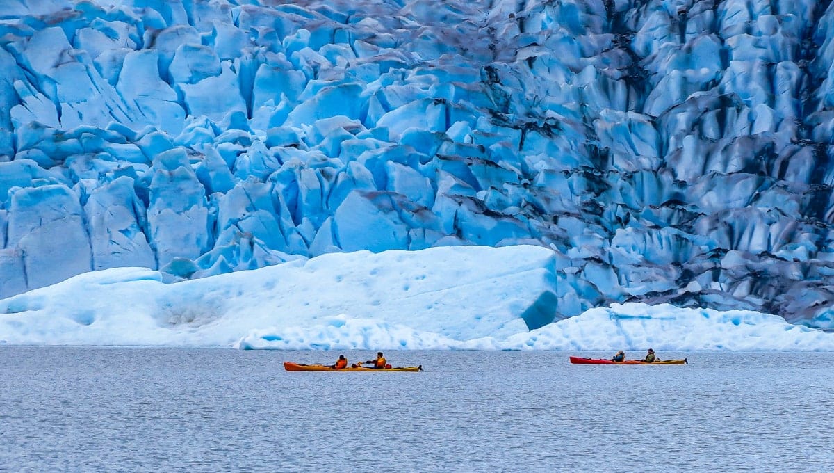 A group of kayakers paddling near the base of a massive blue glacier in Alaska, with jagged ice formations towering above them