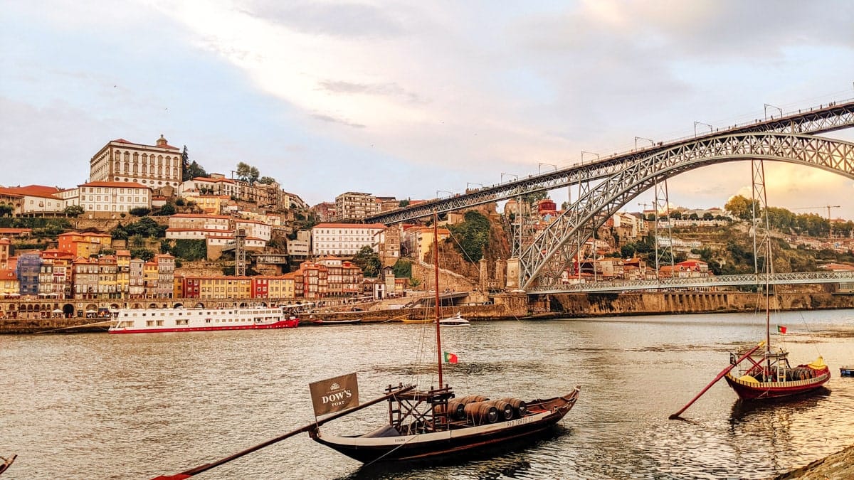 Scenic view of Porto, Portugal – Traditional wooden Rabelo boats floating on the Douro River with the iconic Dom Luís I Bridge and the colorful hillside buildings of Porto in the background.