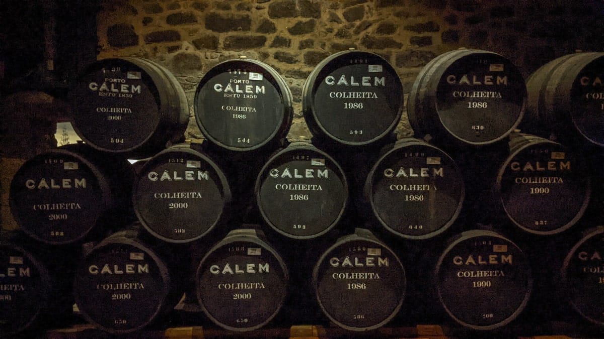 Aging barrels of Port wine – A dimly lit wine cellar with stacked barrels of Calém Port wine, showcasing the rich winemaking heritage of Porto.