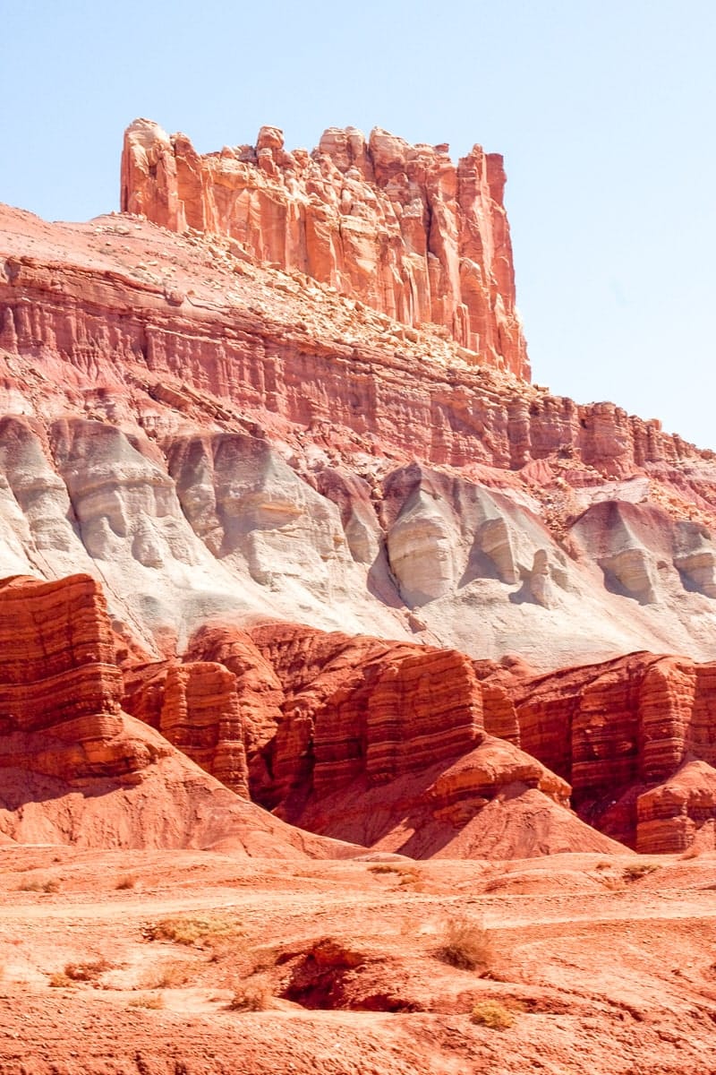 Striking layers of red, orange, white, and gray rock create a dramatic geological formation in Capitol Reef National Park, Utah. The towering cliffs and striped hills tell a story of ancient sediment, with the vibrant hues glowing under bright desert sunlight.