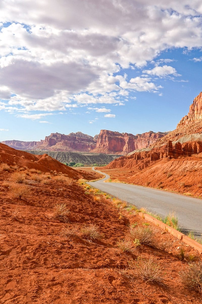 Road winding through Capitol Reef National Park in Utah along the Waterpocket Fold. A winding road leads through the iconic red rock canyons of Capitol Reef National Park, framed by towering cliffs and scattered desert plants. Puffy clouds hang low in the sky, casting soft shadows on the road and creating a perfect setting for a road trip through the Utah desert.